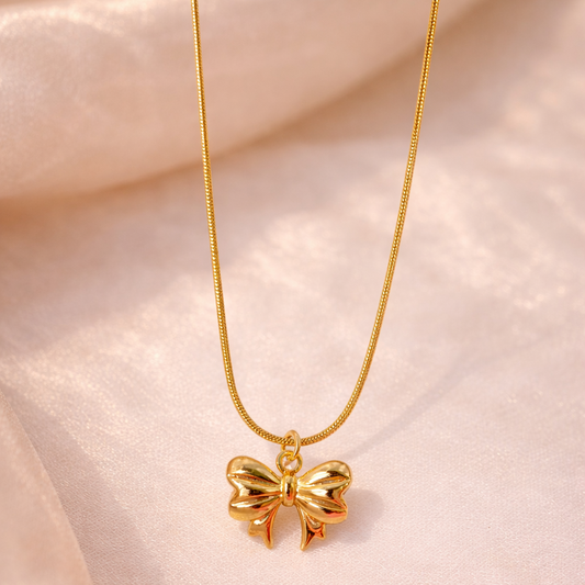 Gold bow-shaped pendant on a chain against a soft pink fabric background