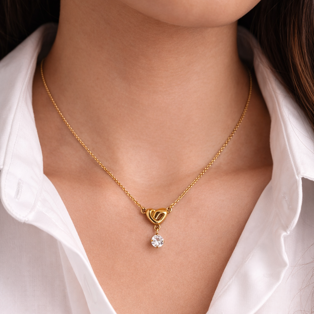 Gold necklace with a heart-shaped pendant and clear gemstone worn by a person in a white shirt.