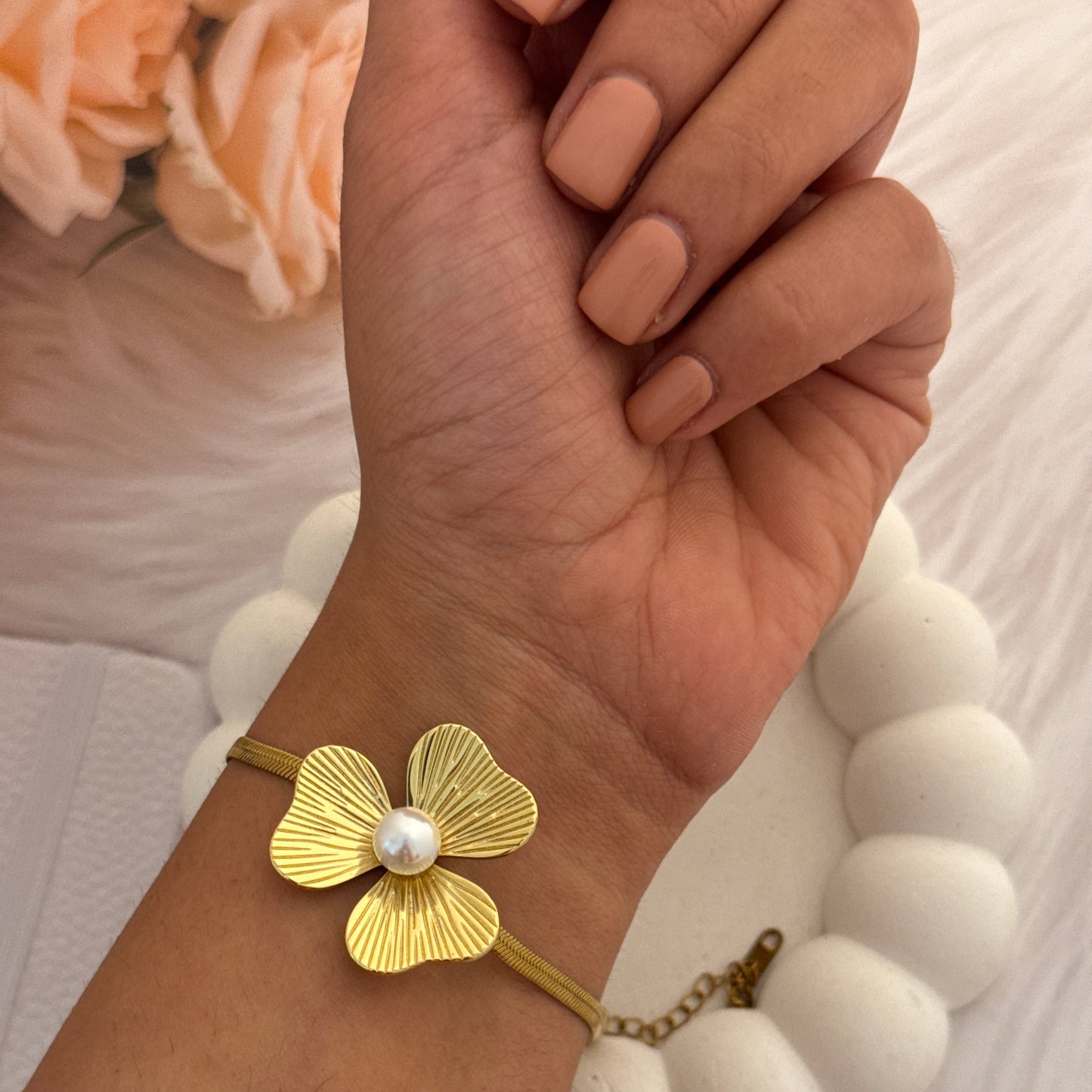 Gold bracelet with a floral design and pearl center on a wrist, against a neutral background.