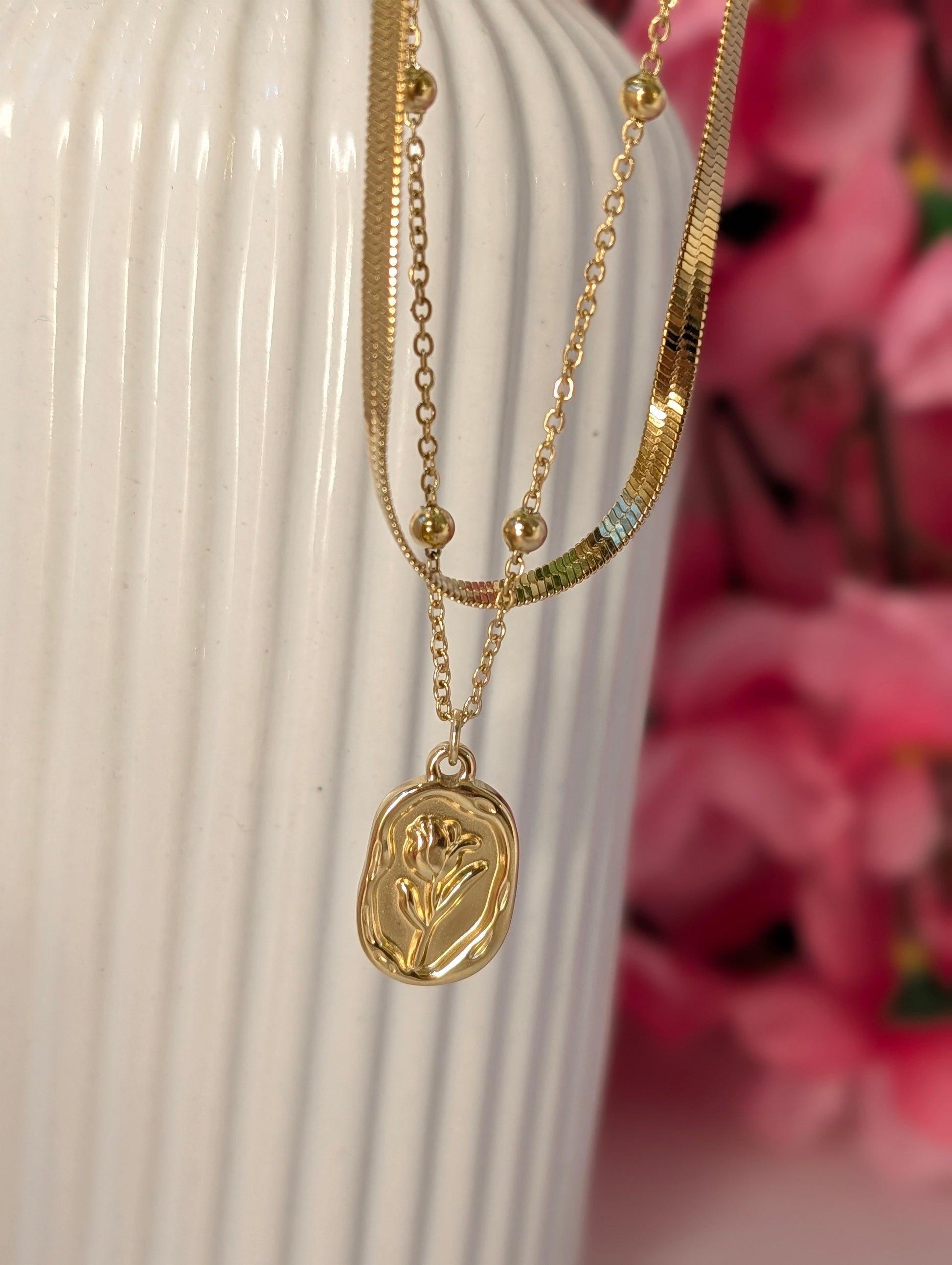Gold necklaces with a pendant on a white surface with pink flowers in the background