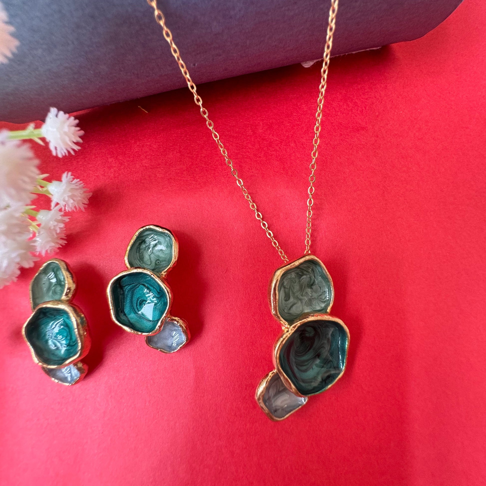 A gold-colored necklace with green emerald-style stones, displayed alongside a matching pair of earrings, against a red background with a floral pattern.
