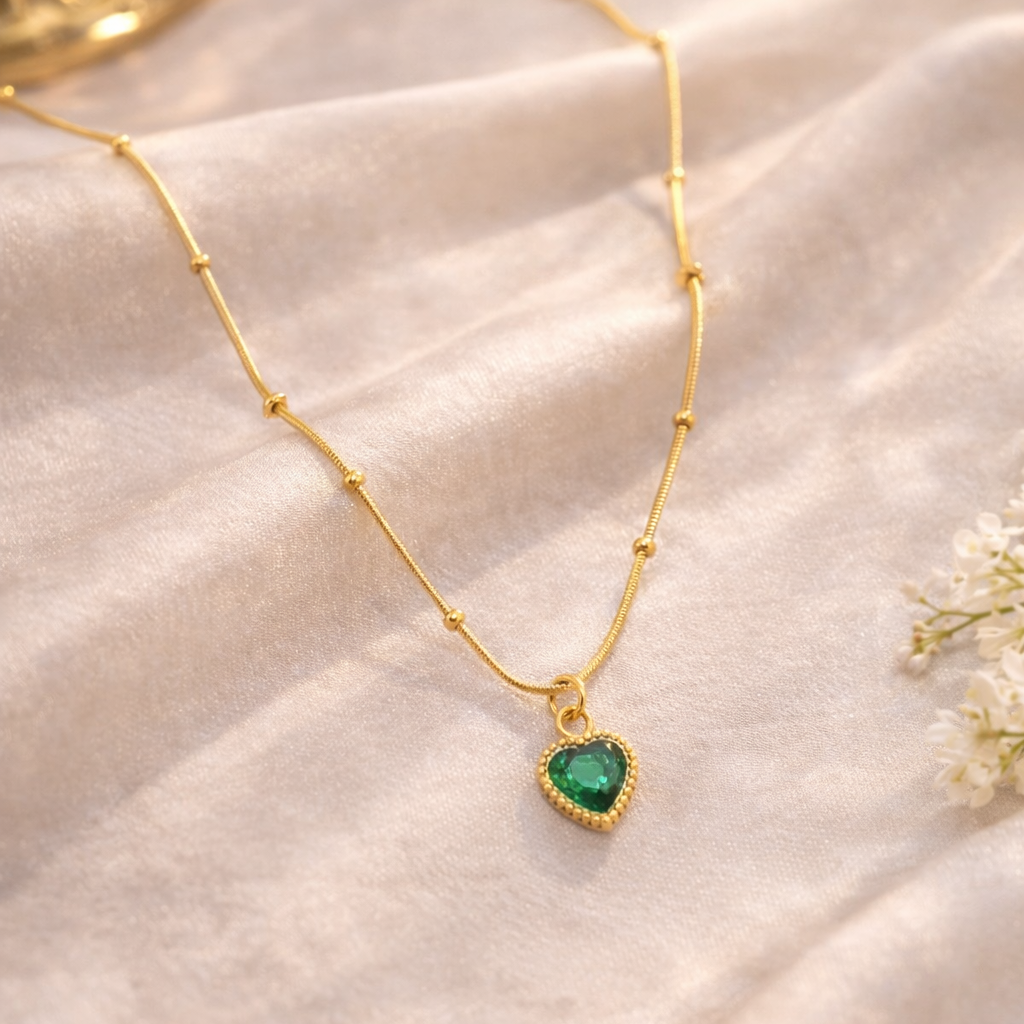 Gold necklace with a heart-shaped green gemstone pendant on a soft beige fabric background.