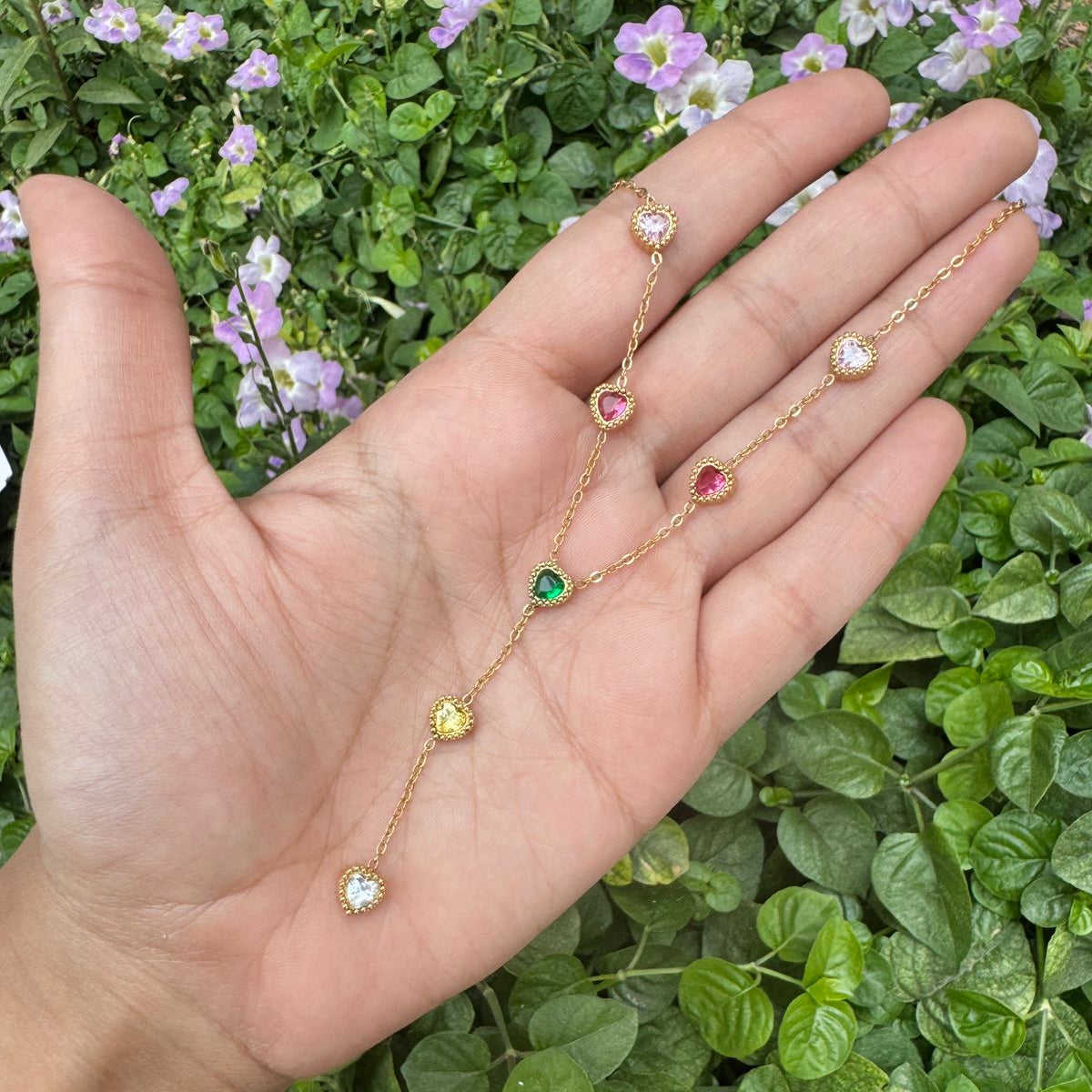 Gold necklace with colorful stones held in a hand against a green leafy background