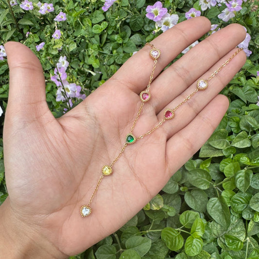 Gold necklace with colorful stones held in a hand against a green leafy background