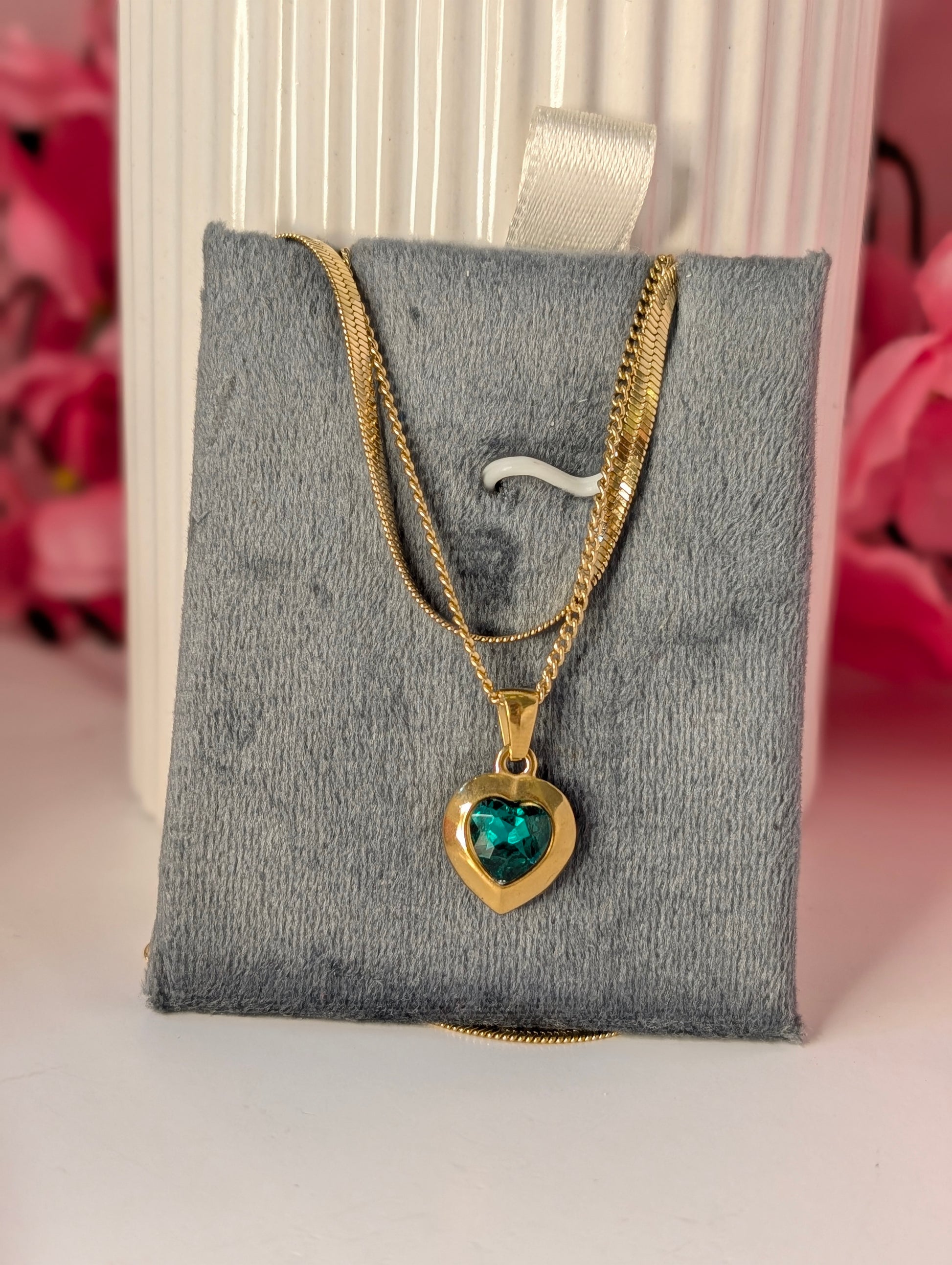 Gold necklace with a heart-shaped pendant on a gray cushion, pink flowers in the background