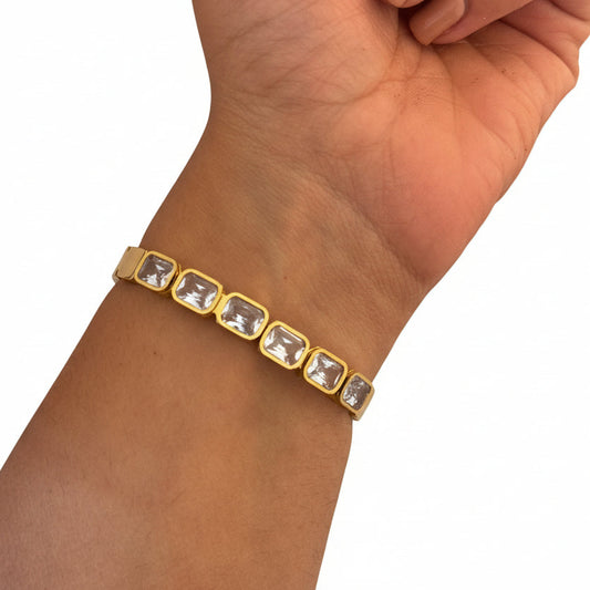 Gold bracelet with square stones on a wrist, with a blurred background of flowers and fabric.