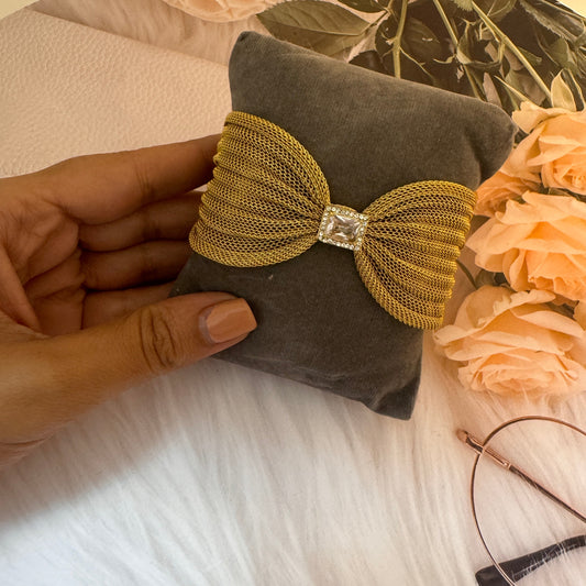 Hand holding a decorative item with a gold bow and gray fabric, with flowers and a necklace in the background.