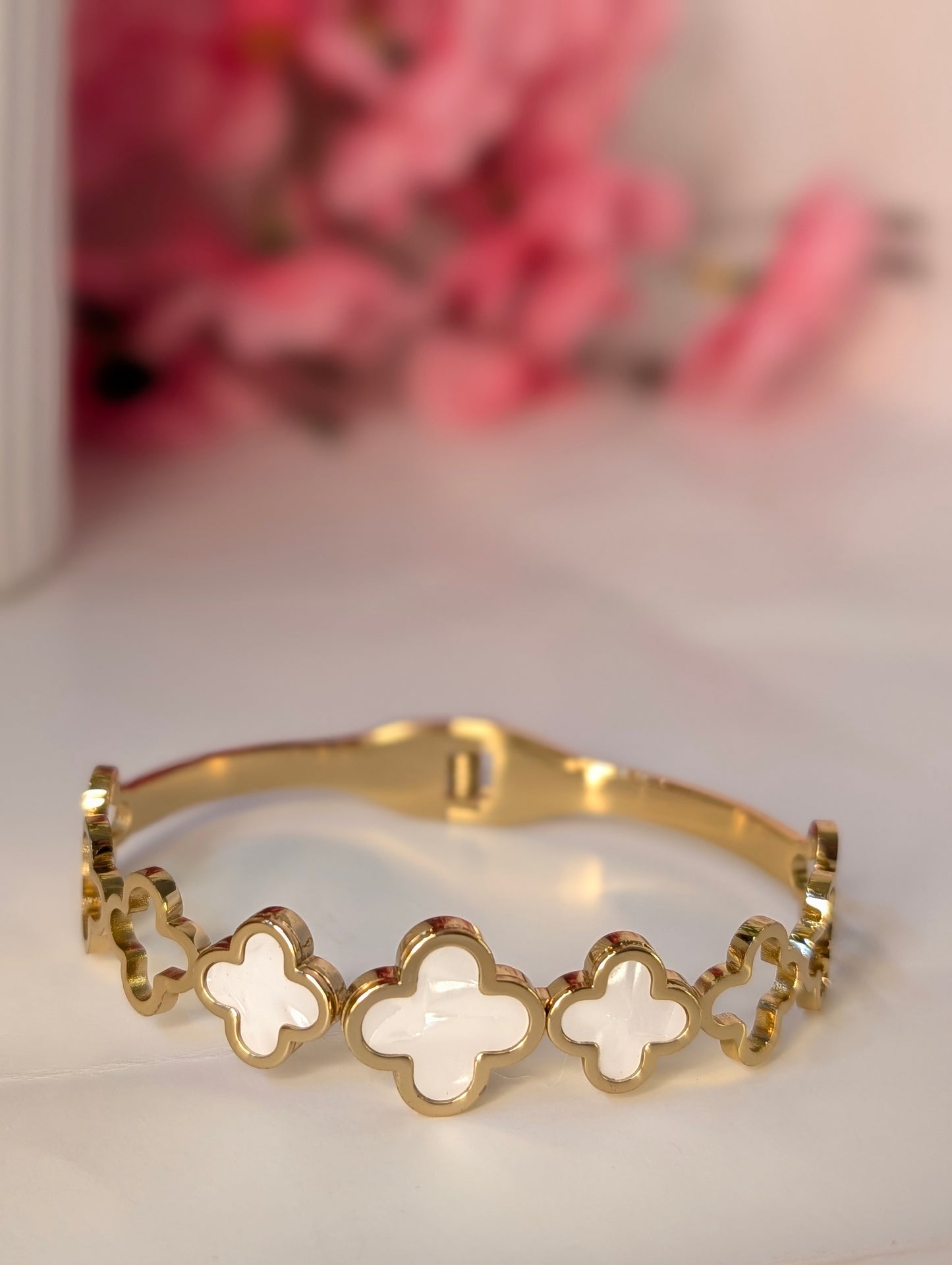Gold bracelet with white clover-shaped stones on a light surface with pink flowers in the background