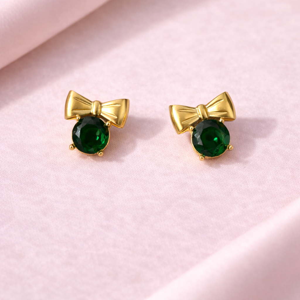 Gold bow-shaped earrings with green gemstones on a pink background
