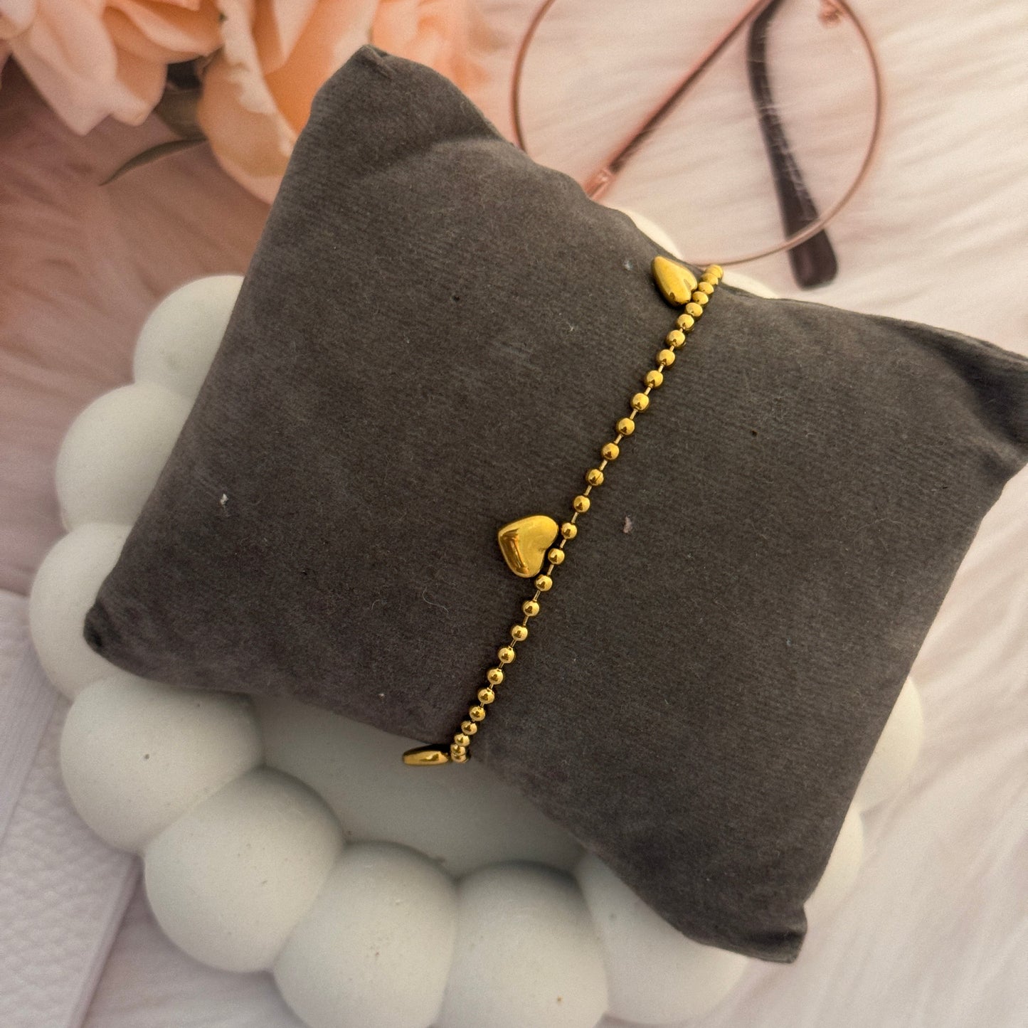 Gold bracelet with heart charms on a gray pillow surrounded by flowers and glasses.