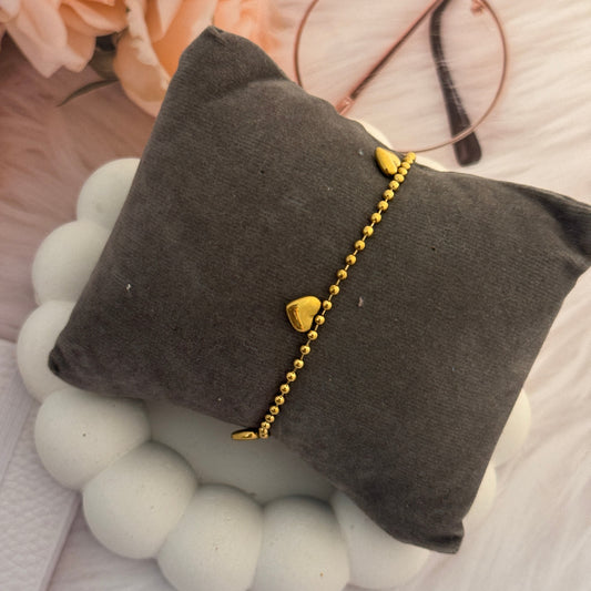 Gold bracelet with heart charms on a gray pillow surrounded by flowers and glasses.