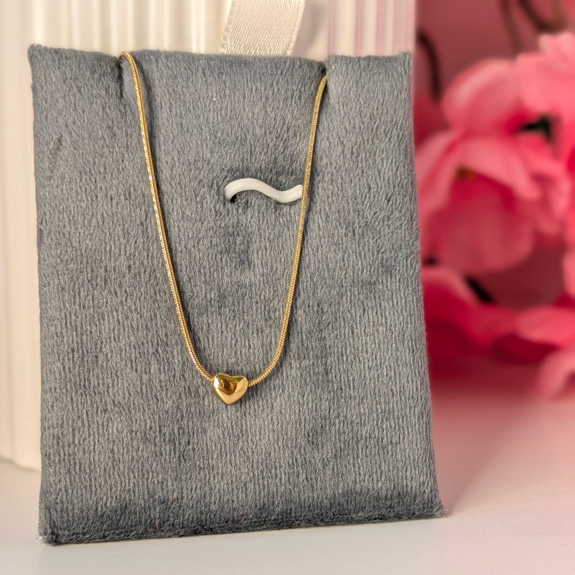 Gold necklace with a heart pendant on a gray jewelry display pillow against a pink floral background