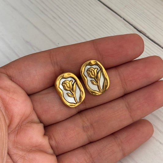 Gold and white earrings held in a hand on a light wooden surface