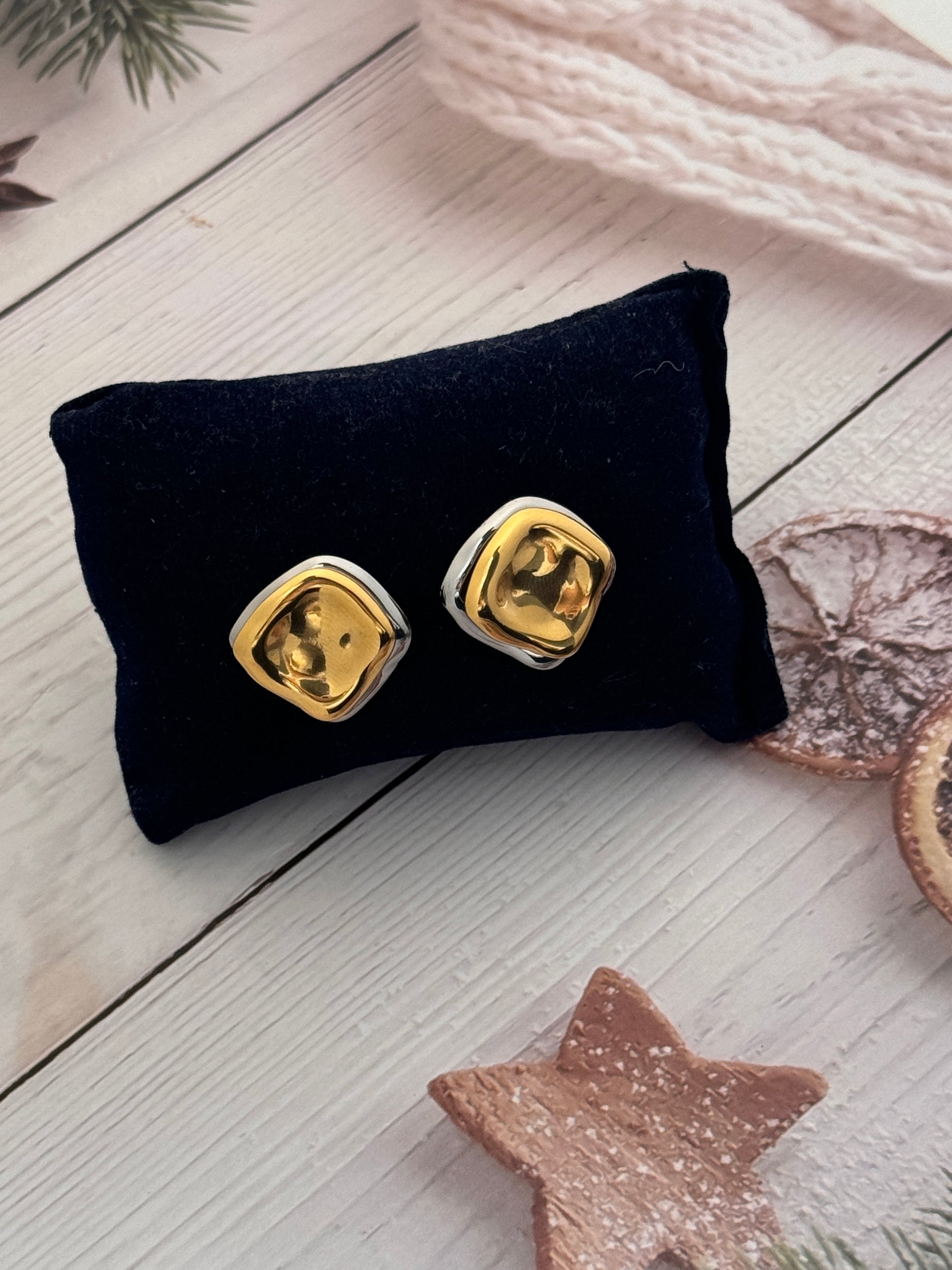 Gold heart-shaped earrings on a black pillow with a white wooden surface and decorative items in the background.