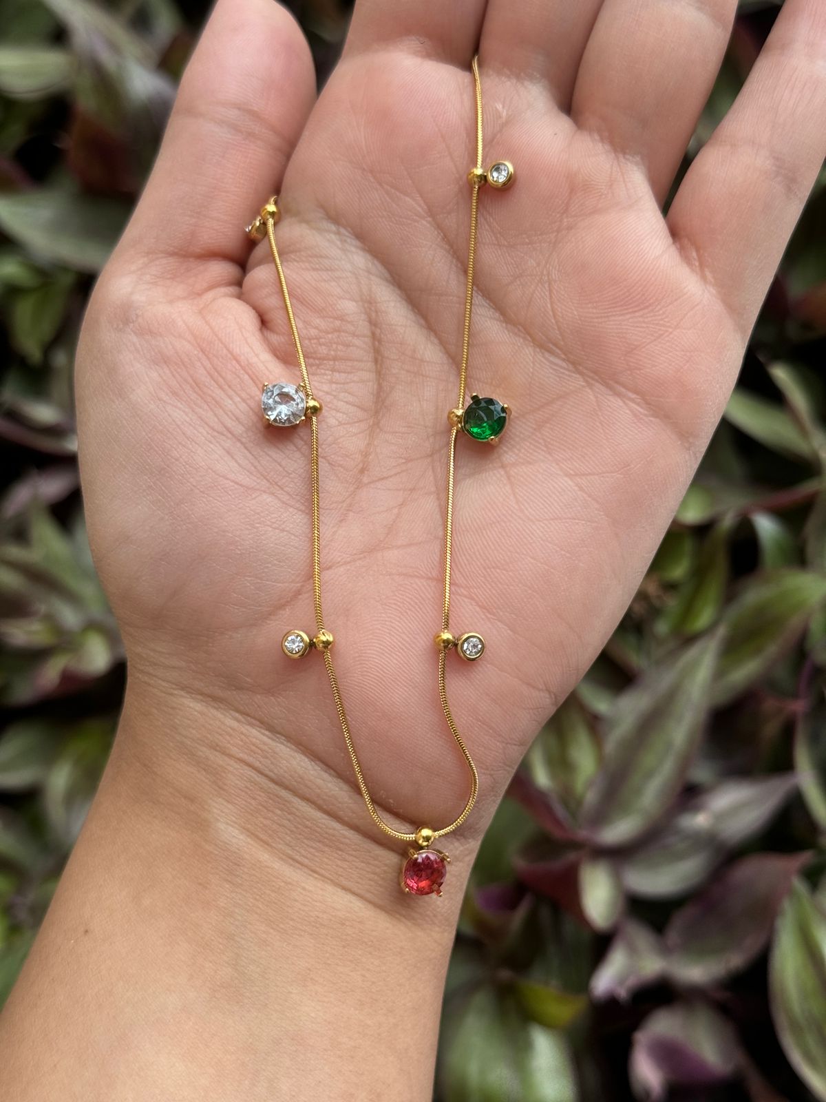 Gold necklace with gemstone pendants held in a hand against a blurred plant background
