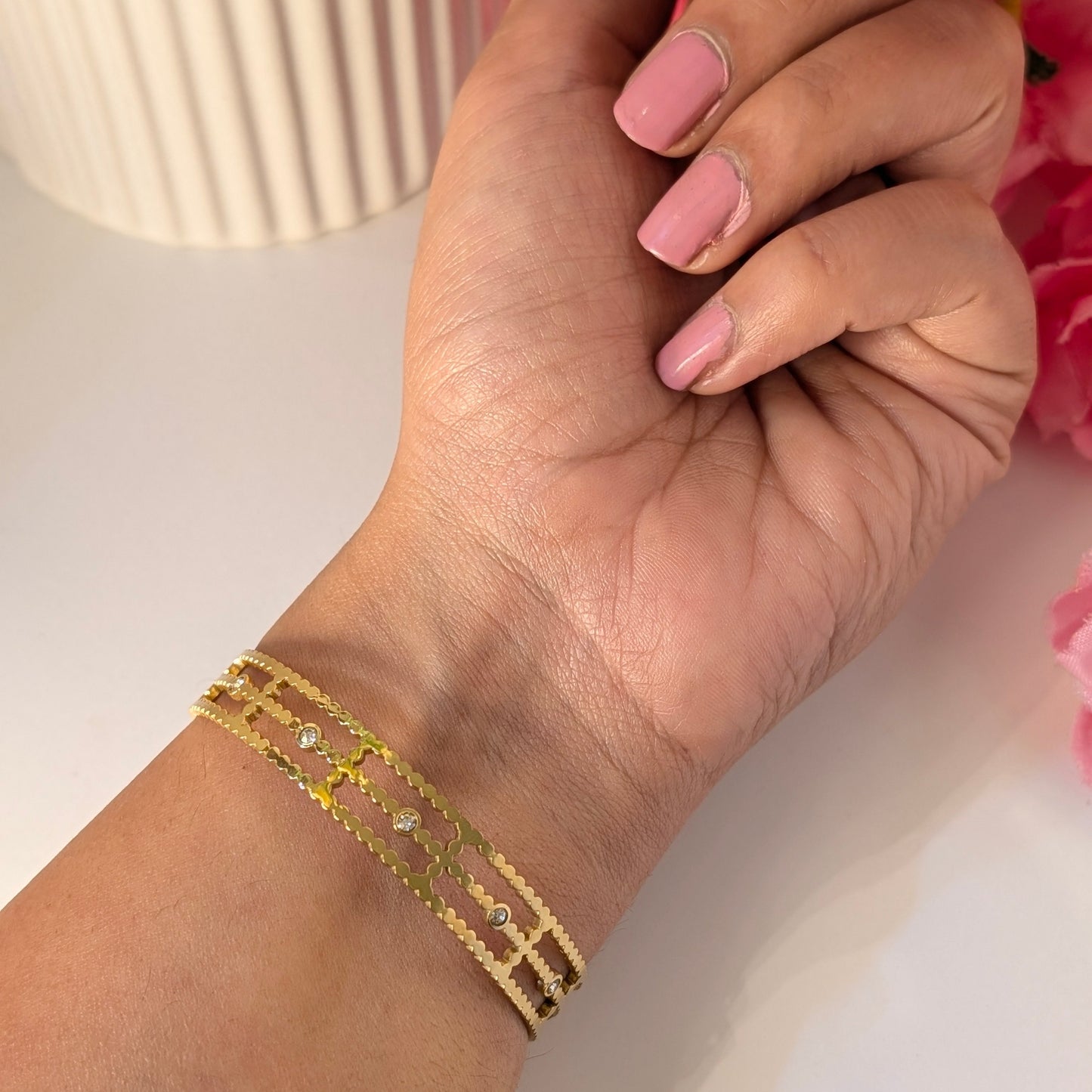 Hand wearing a gold bracelet with pink flowers and a white vase in the background