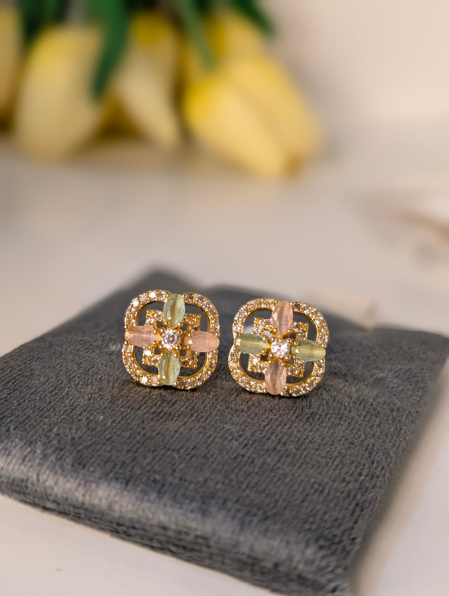 Gold and green earrings on a gray surface with blurred yellow flowers in the background