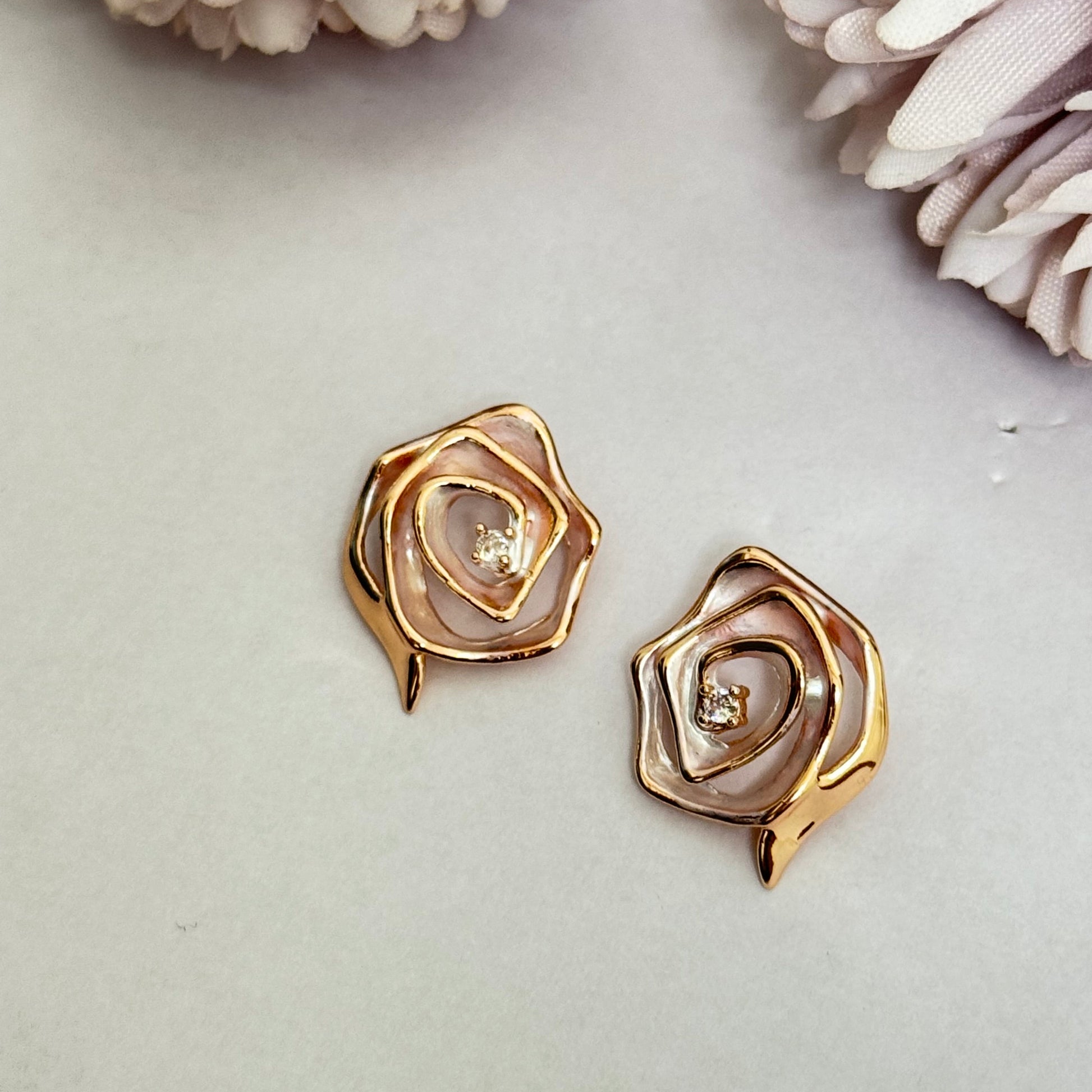 A pair of rose-shaped stud earrings with a gold-tone finish on a white background with pale pink flowers.