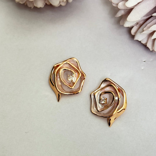 A pair of rose-shaped stud earrings with a gold-tone finish on a white background with pale pink flowers.