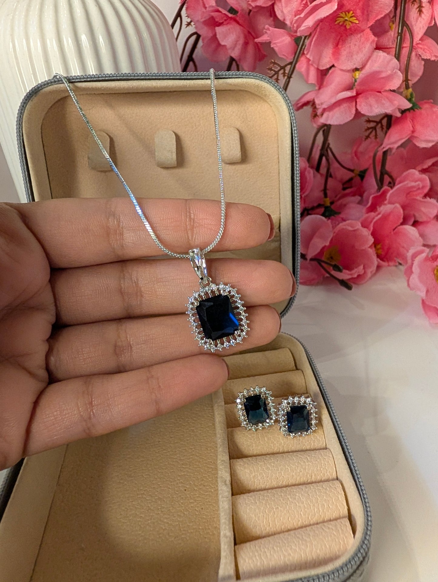 Silver necklace with a blue pendant held over an open jewelry box with earrings inside, pink flowers in the background.
