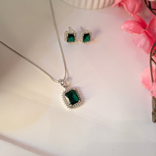 Silver necklace with green gemstone and matching earrings on a white surface with pink flowers.