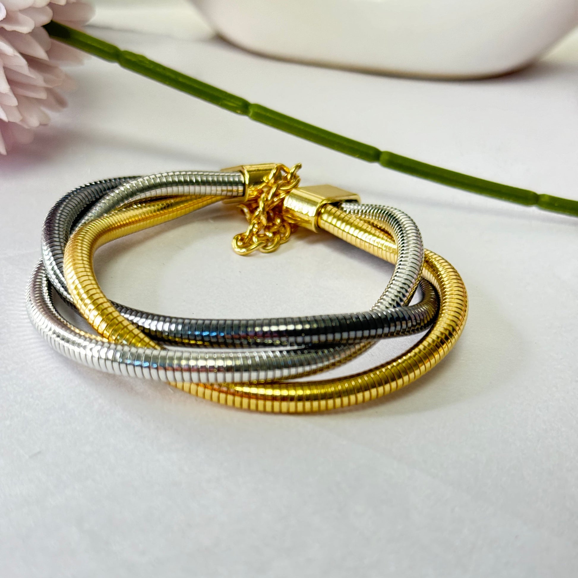 A two-tone golden and silver layered bracelet with a decorative element in the center, placed on a white surface next to a flower and a white cup.