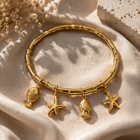 Gold bracelet with starfish, fish, and shell charms on a beige surface with dried flowers.
