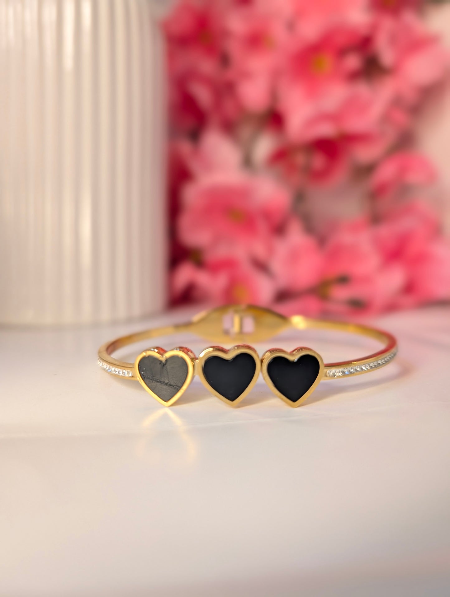 Gold bracelet with three black heart charms on a white surface with pink flowers in the background