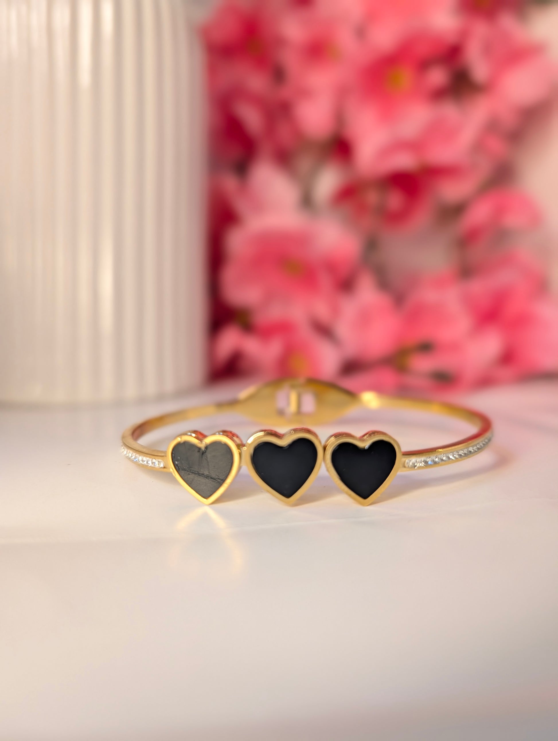 Gold bracelet with three black heart charms on a white surface with pink flowers in the background