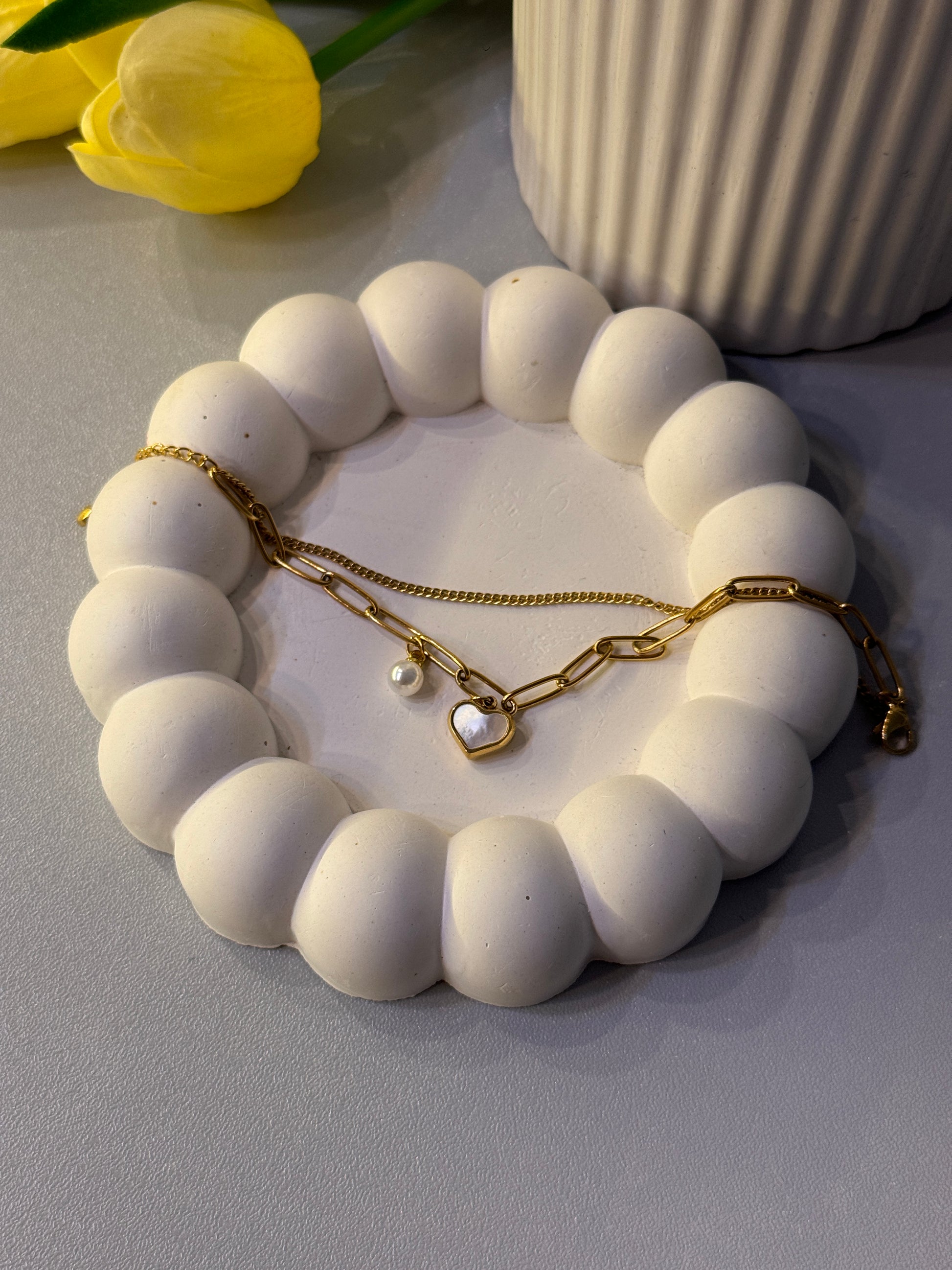 White beaded bracelet on a white stand with a gold chain and heart charm, against a light gray background.