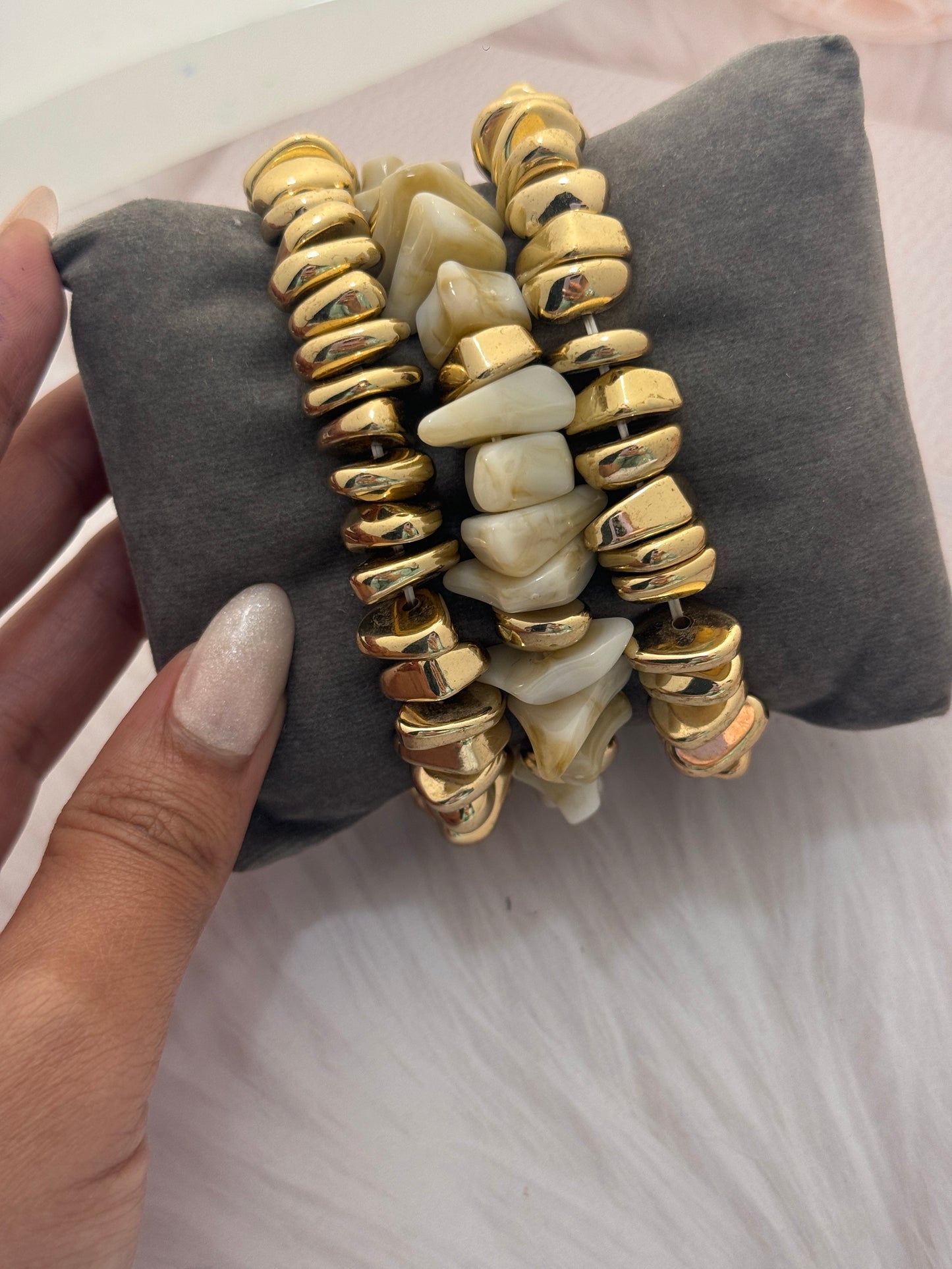 Stack of gold and beige beaded bracelets on a gray pillow with a hand holding them.
