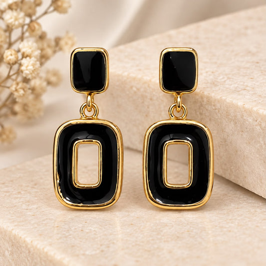 A pair of gold and black geometric drop earrings displayed on a beige stone block with dried flowers in the background.