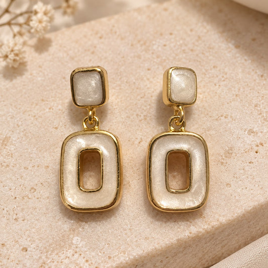 A pair of gold-tone drop earrings with rectangular mother-of-pearl inlays, displayed on a beige stone surface with dried flowers in the background.