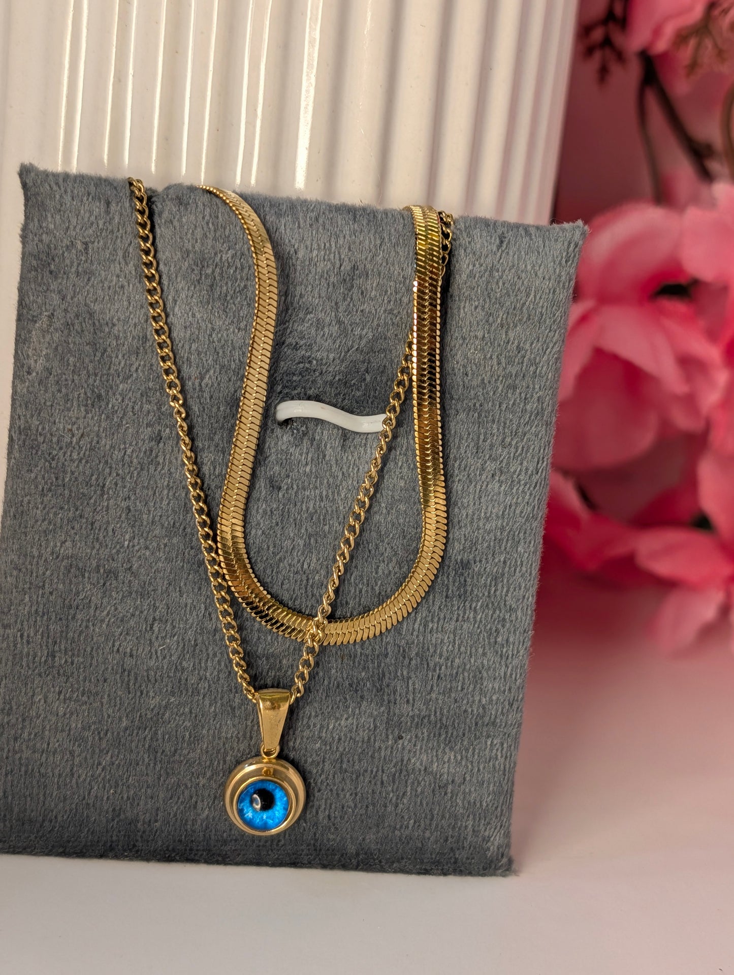 Gold necklace with a blue pendant on a gray stand with pink flowers in the background