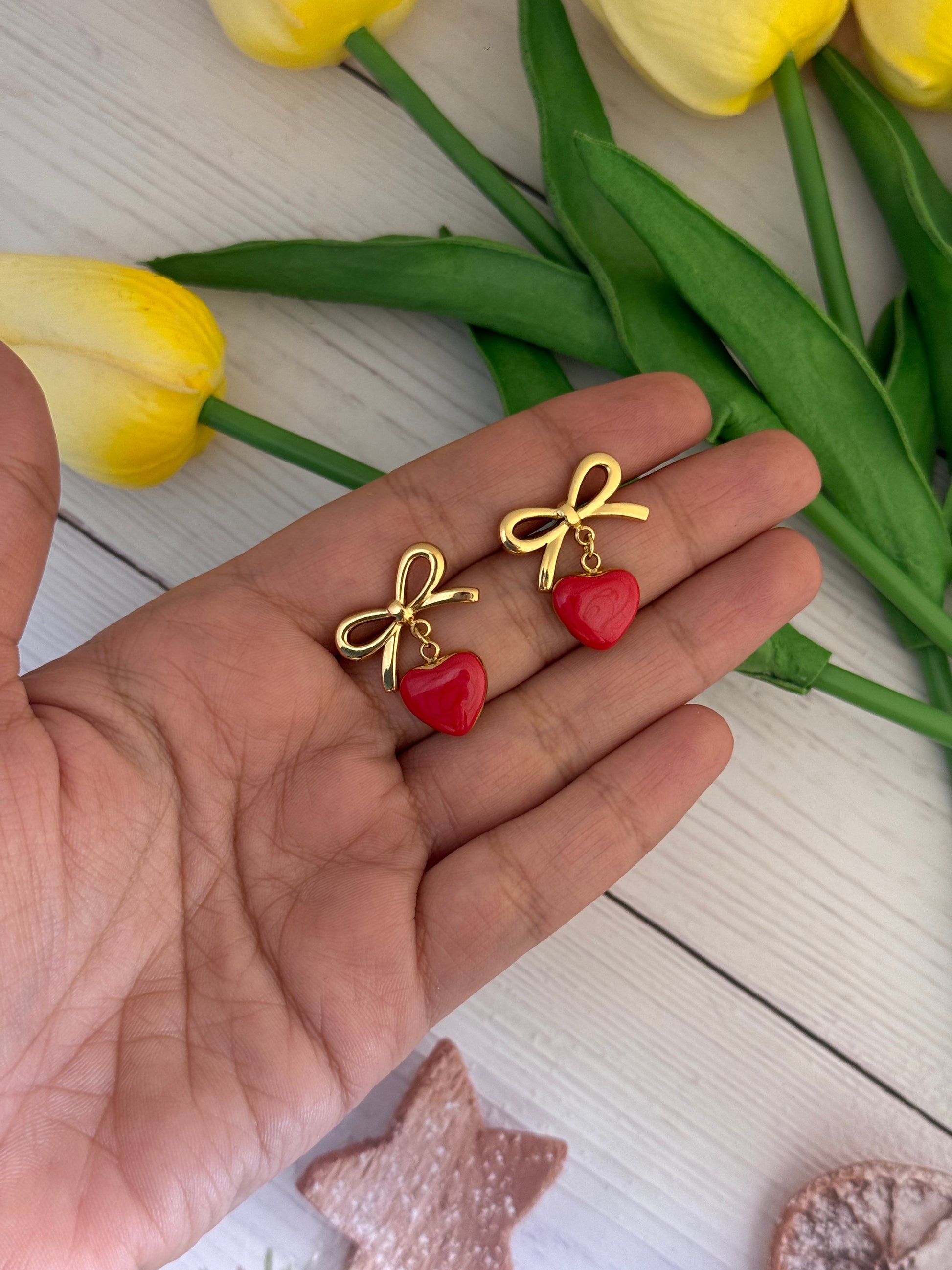 Hand holding two small gold bow-shaped earrings with red centers, surrounded by yellow tulips on a light wooden surface.