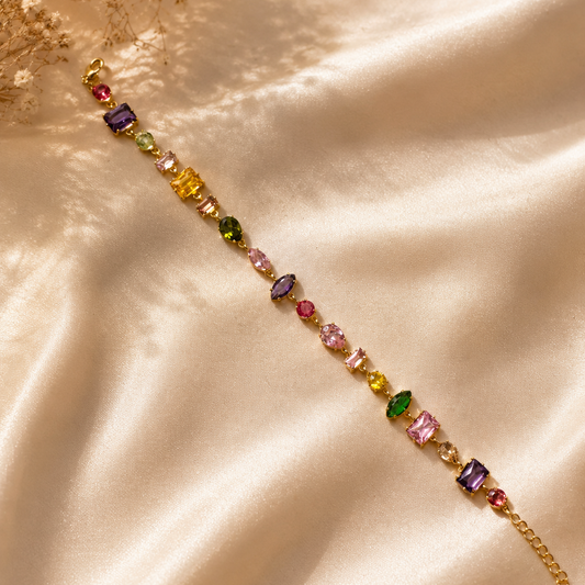 A gold chain bracelet with colorful gemstone accents, laid diagonally on draped champagne satin fabric.