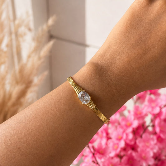 A gold-toned bangle bracelet with a central oval clear gemstone, worn on a person's wrist against a softly blurred background with pink flowers.