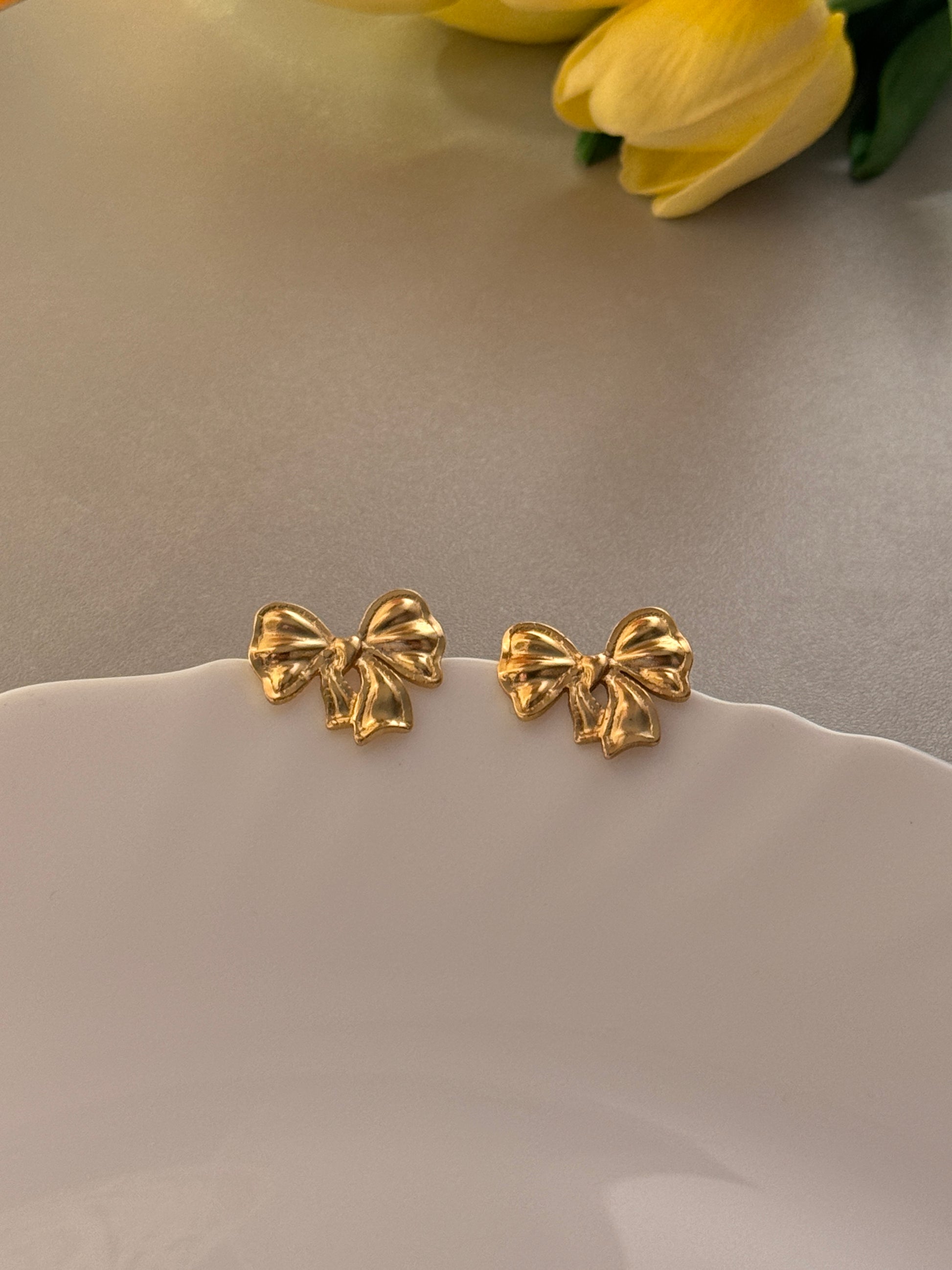 Gold bow-shaped earrings on a light gray background with a yellow flower.