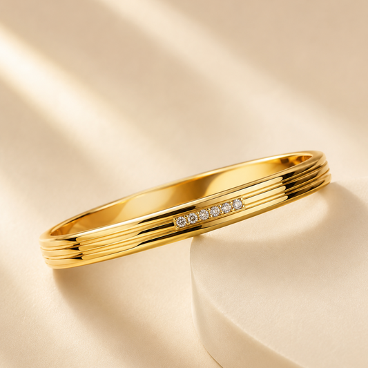 A polished gold bangle bracelet with a row of small clear stones, displayed on a cream-colored pedestal with soft diagonal shadows.