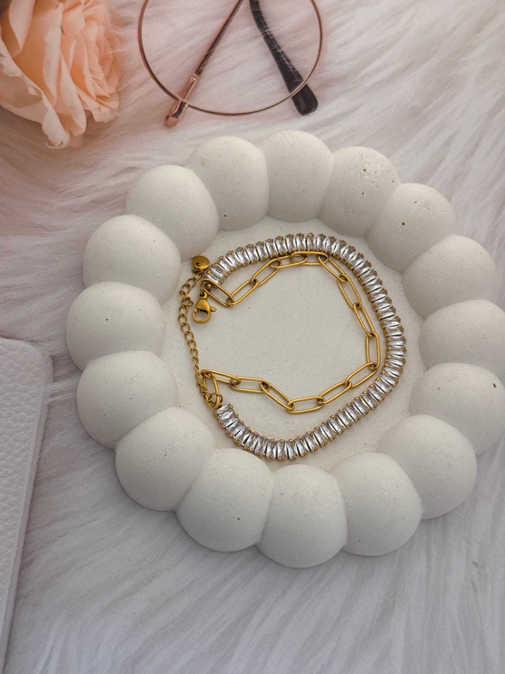 Gold and silver chain necklace on a white stone surface with a pink flower and glasses in the background.