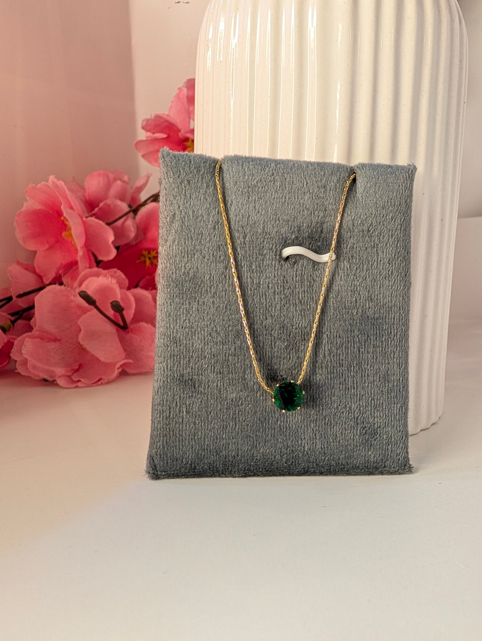 Necklace with a green pendant on a gray jewelry pillow, with pink flowers in the background.