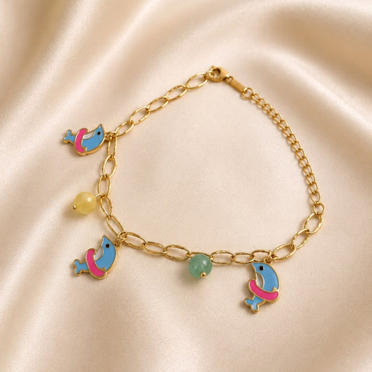 A gold chain bracelet featuring four colorful dolphin charms in blue, pink, and light green, displayed on a draped beige satin fabric.