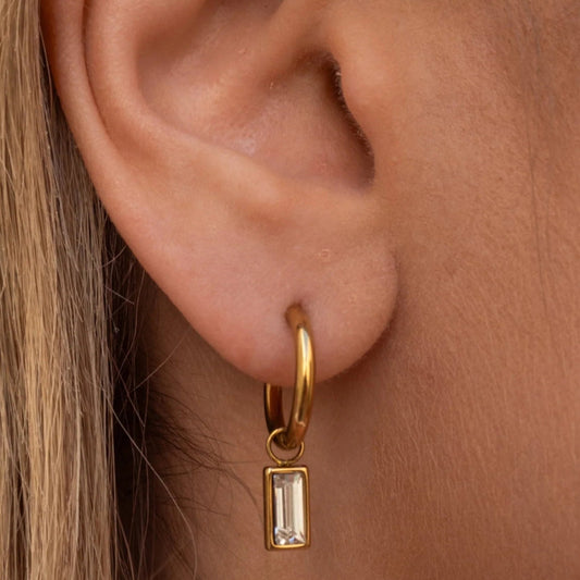 Gold hoop earring with a rectangular pendant worn on an ear.