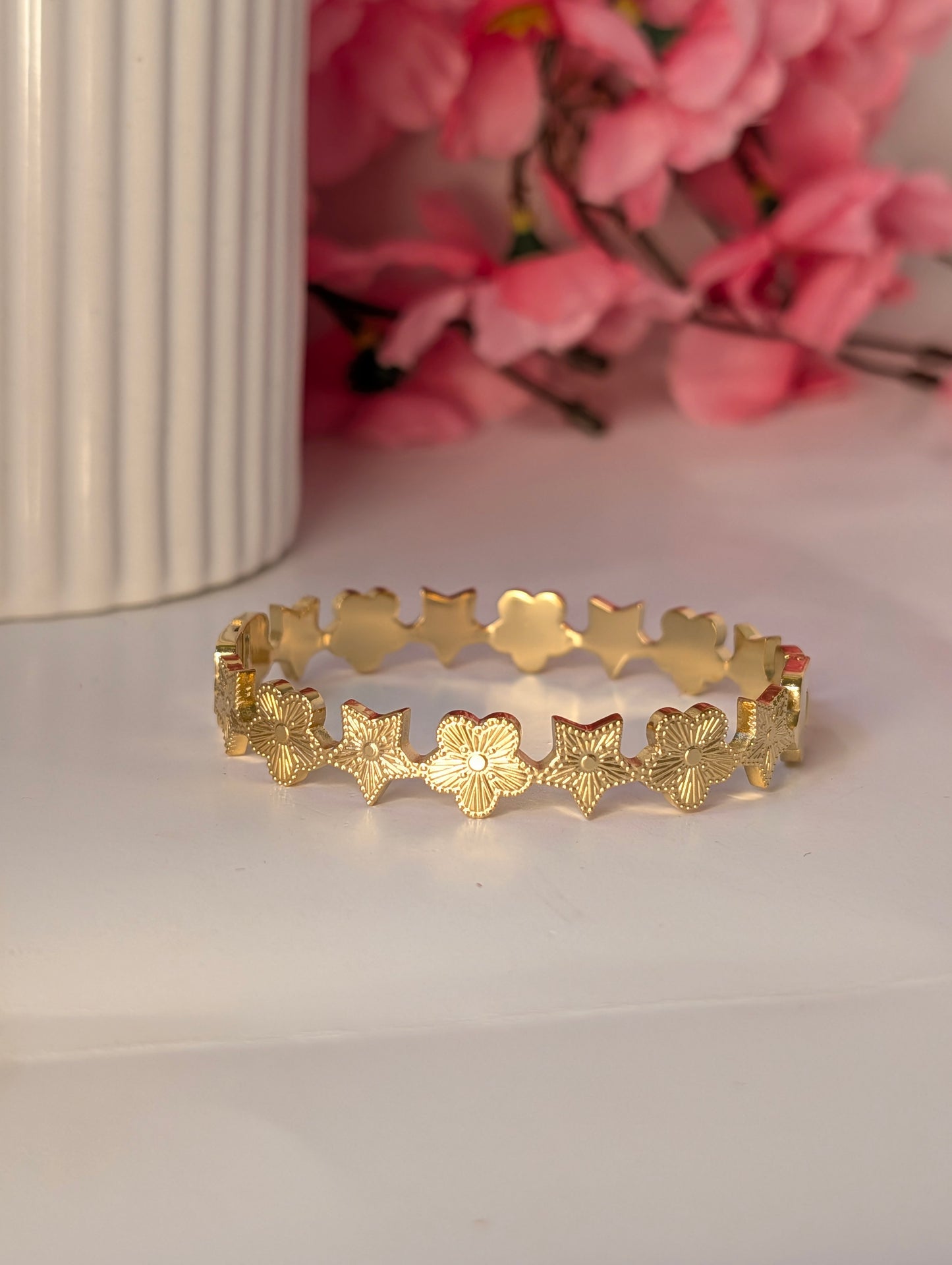 Gold bracelet with star and floral patterns on a white surface with pink flowers in the background