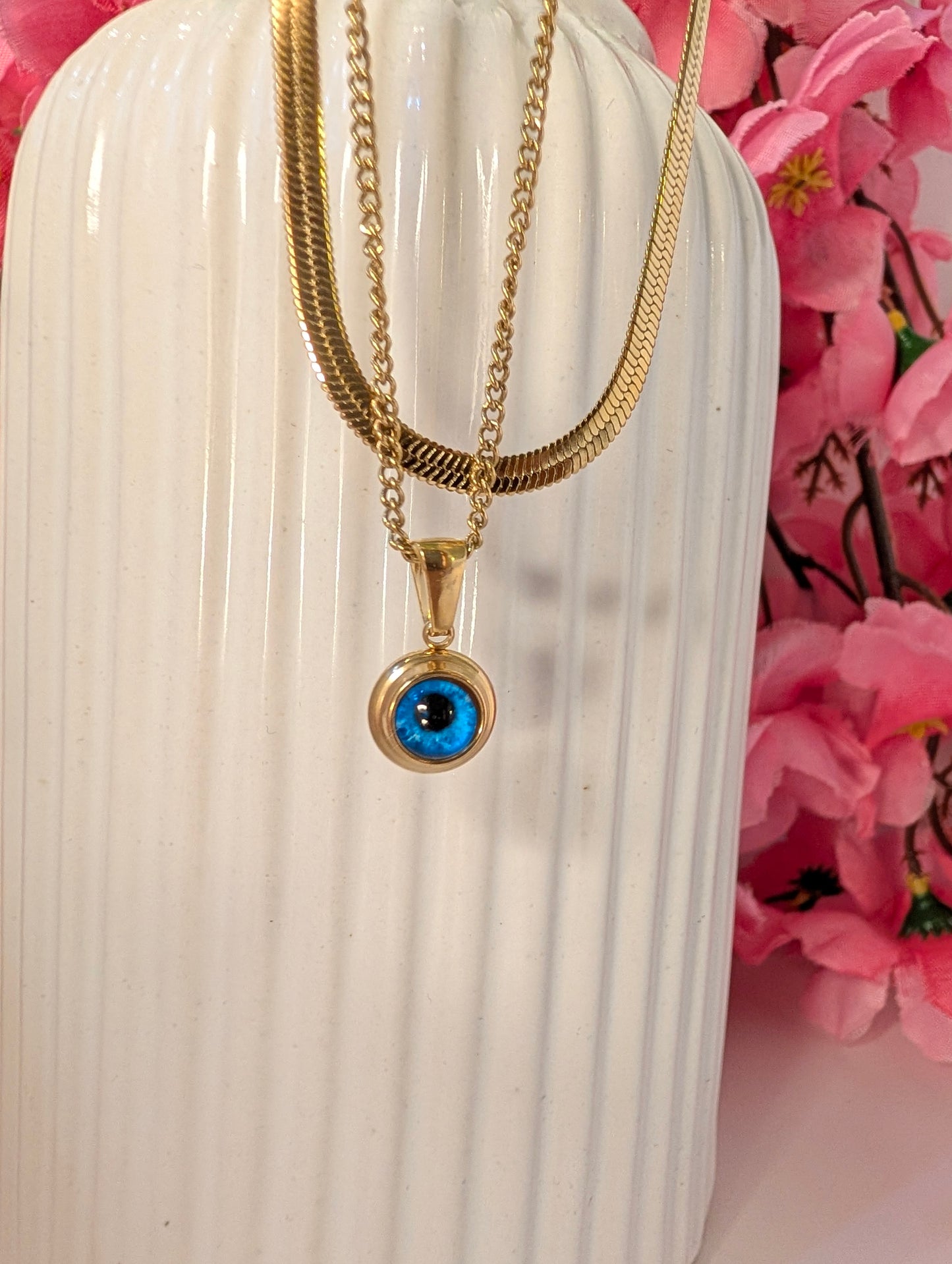 Gold necklace with a blue eye pendant on a white vase with pink flowers in the background