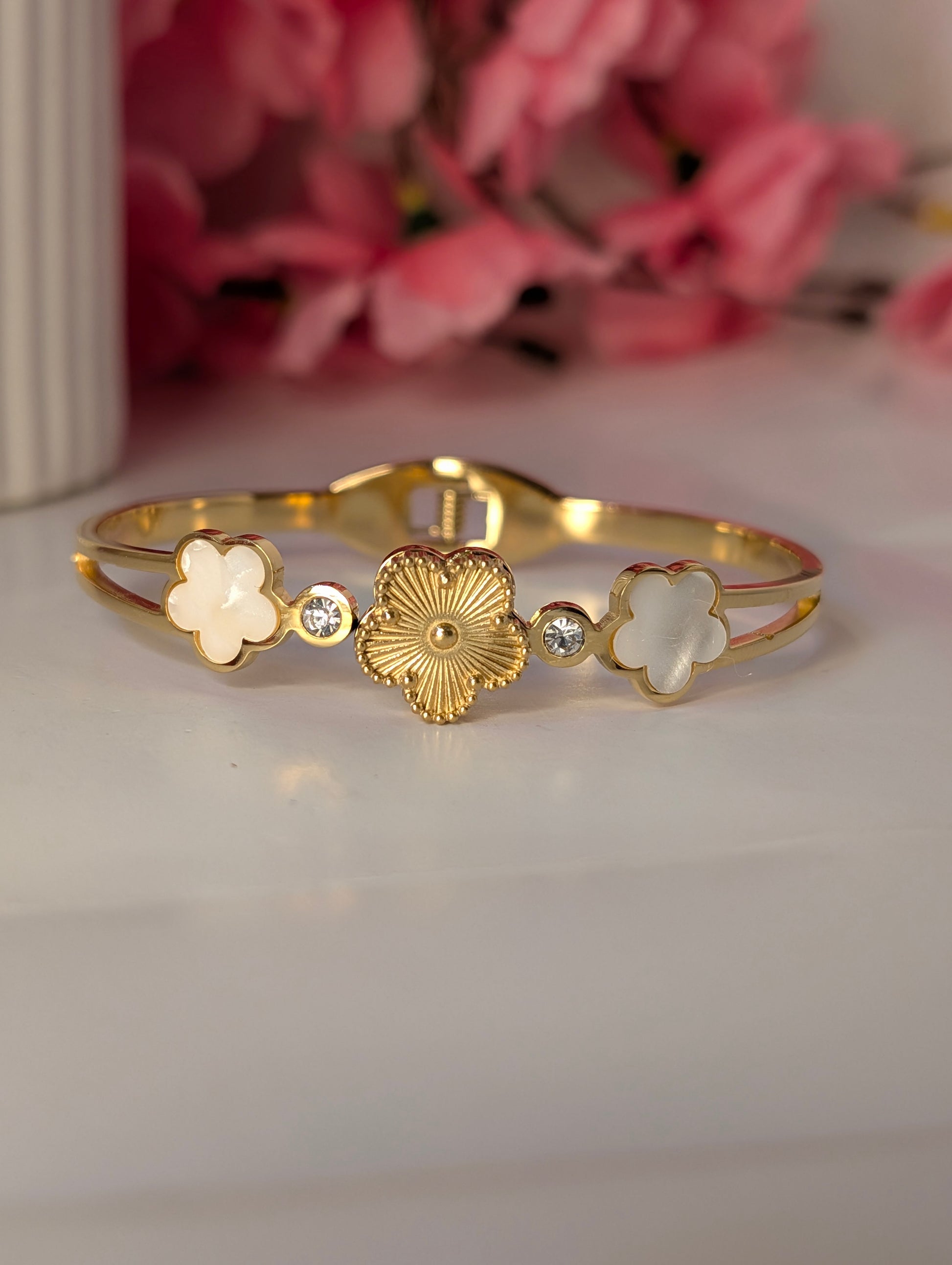 Gold bracelet with floral and heart designs on a white surface with pink flowers in the background