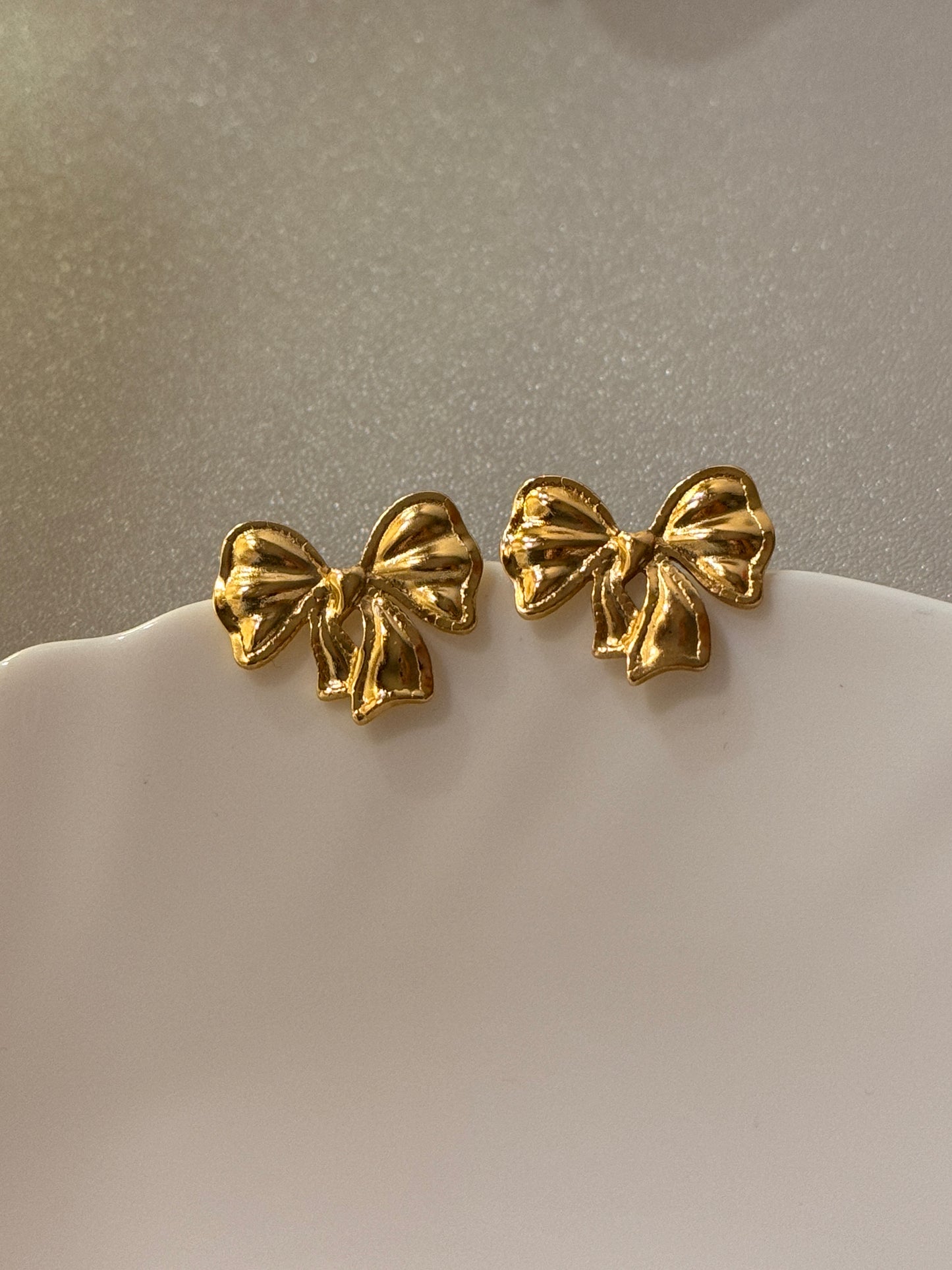 Gold bow-shaped earrings on a white ear model against a gray background