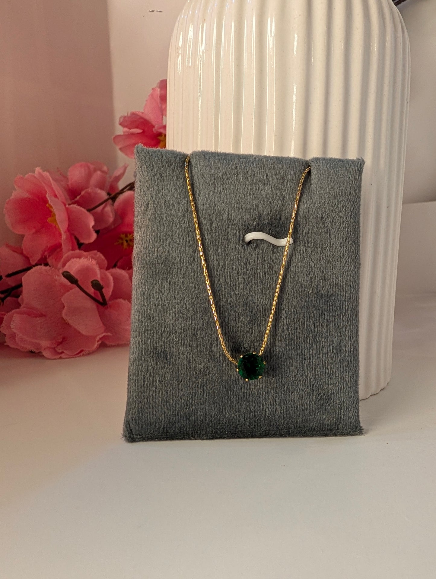 Necklace with a green pendant on a gray jewelry pillow, with pink flowers and a white vase in the background.