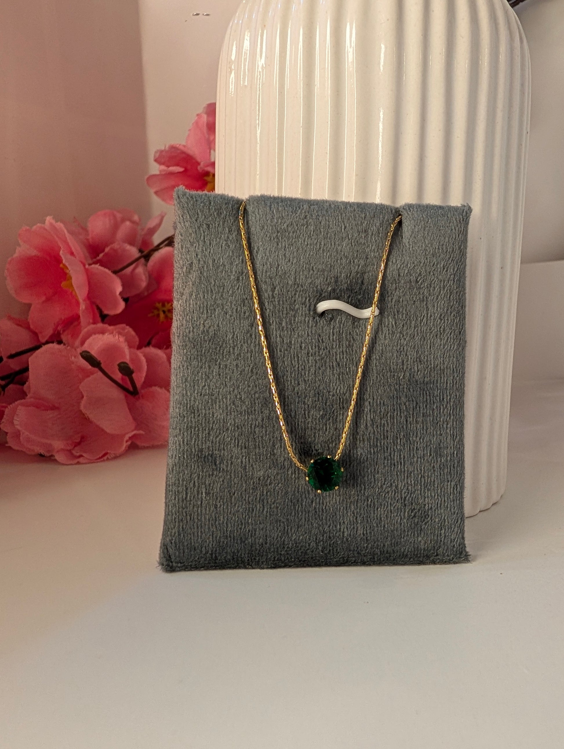 Necklace with a green pendant on a gray jewelry pillow, with pink flowers and a white vase in the background.