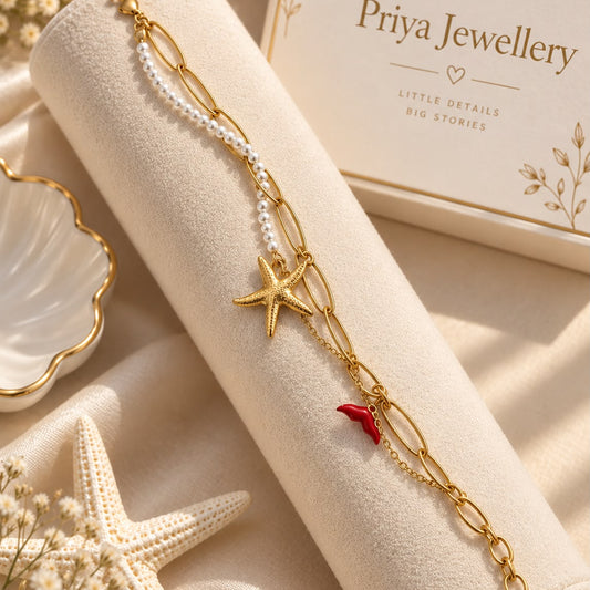 A gold chain bracelet with a starfish charm, a small red coral accent, and a strand of white faux pearls, displayed on a cream jewelry roll. The background includes a starfish prop, dried flowers, and a branded 'Priya Jewellery' box.